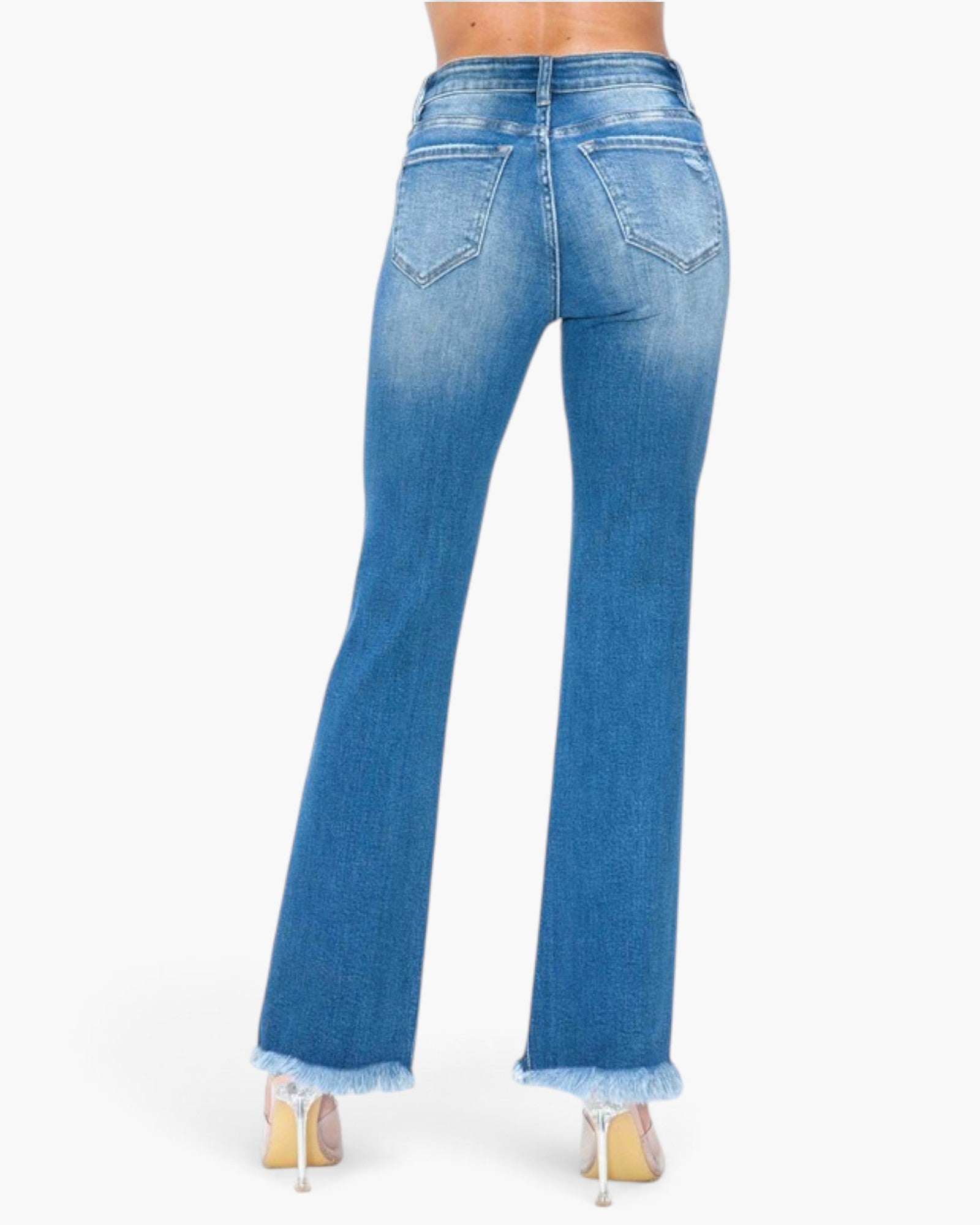 Rear view of a person wearing blue flare jeans with a frayed hem and clear high heels, shown against a plain light gray studio background.