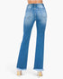Rear view of a person wearing blue flare jeans with a frayed hem and clear high heels, shown against a plain light gray studio background.