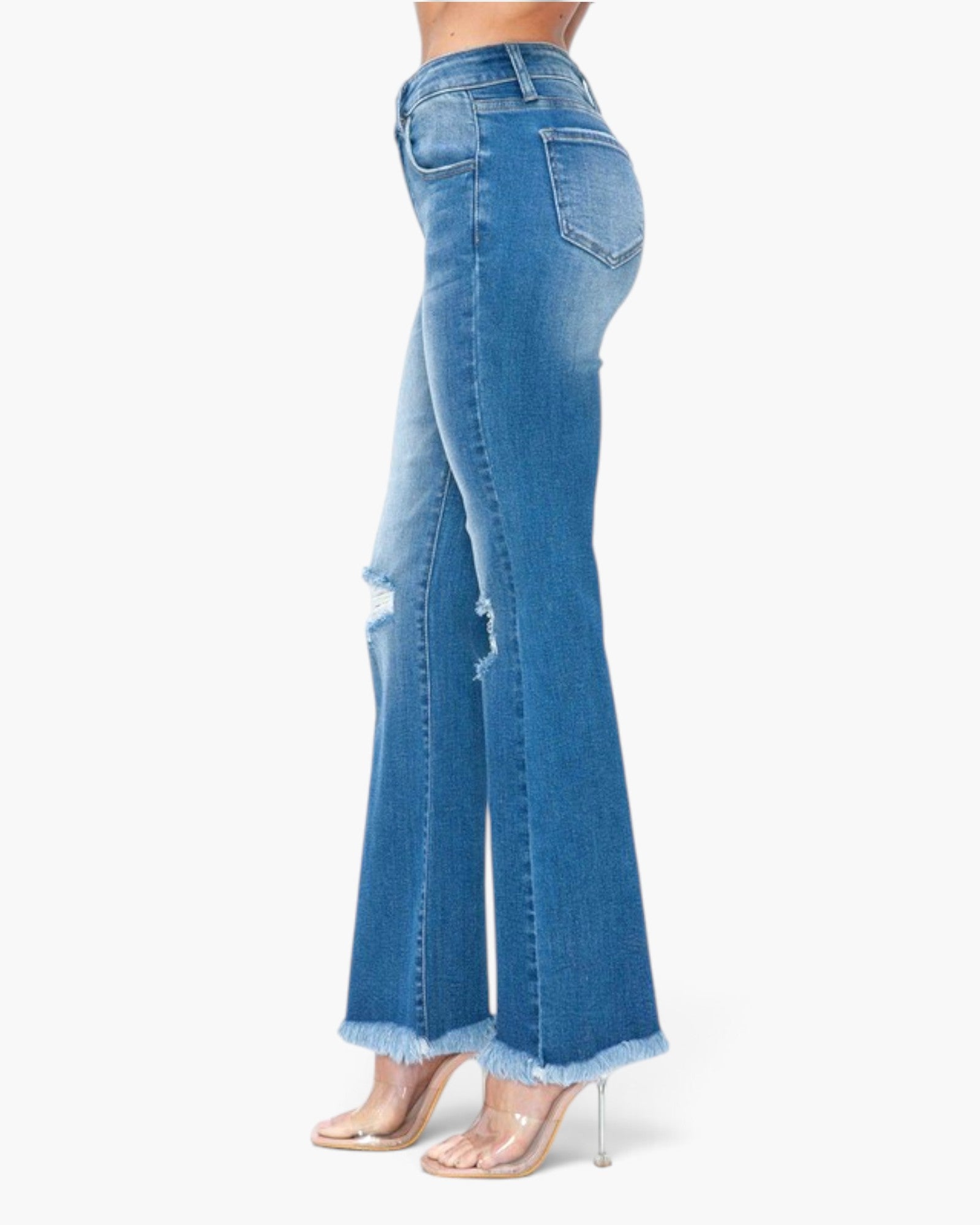 Side view of a model wearing medium-wash blue denim flare jeans with distressed rips and frayed hems, paired with clear high-heeled sandals, against a plain white background.
