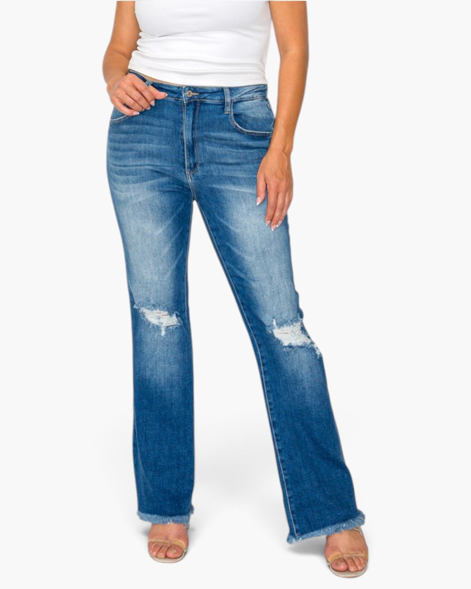 A model wearing a pair of medium-wash blue denim jeans with a distressed, slightly flared leg, paired with a white tank top and beige sandals, against a plain white background.