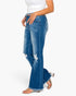 A model wearing a pair of medium-wash blue denim jeans with a flared leg, frayed hems, and distressed details on the knees, paired with a white top and clear block-heel sandals against a plain white background.
