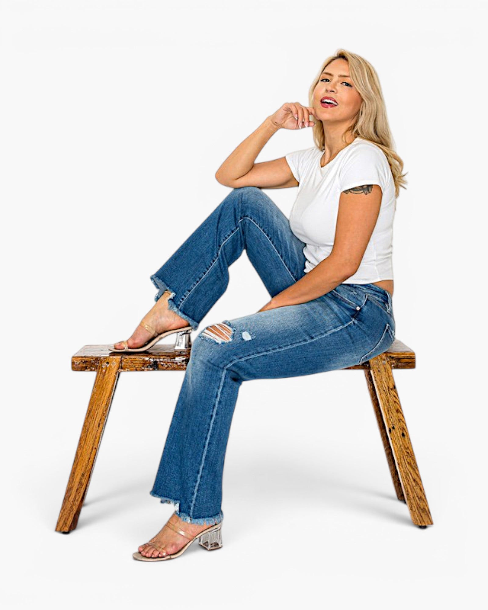 A woman sits on a rustic wooden bench wearing a white t-shirt, blue distressed flare jeans, and clear heeled sandals against a plain white studio background.
