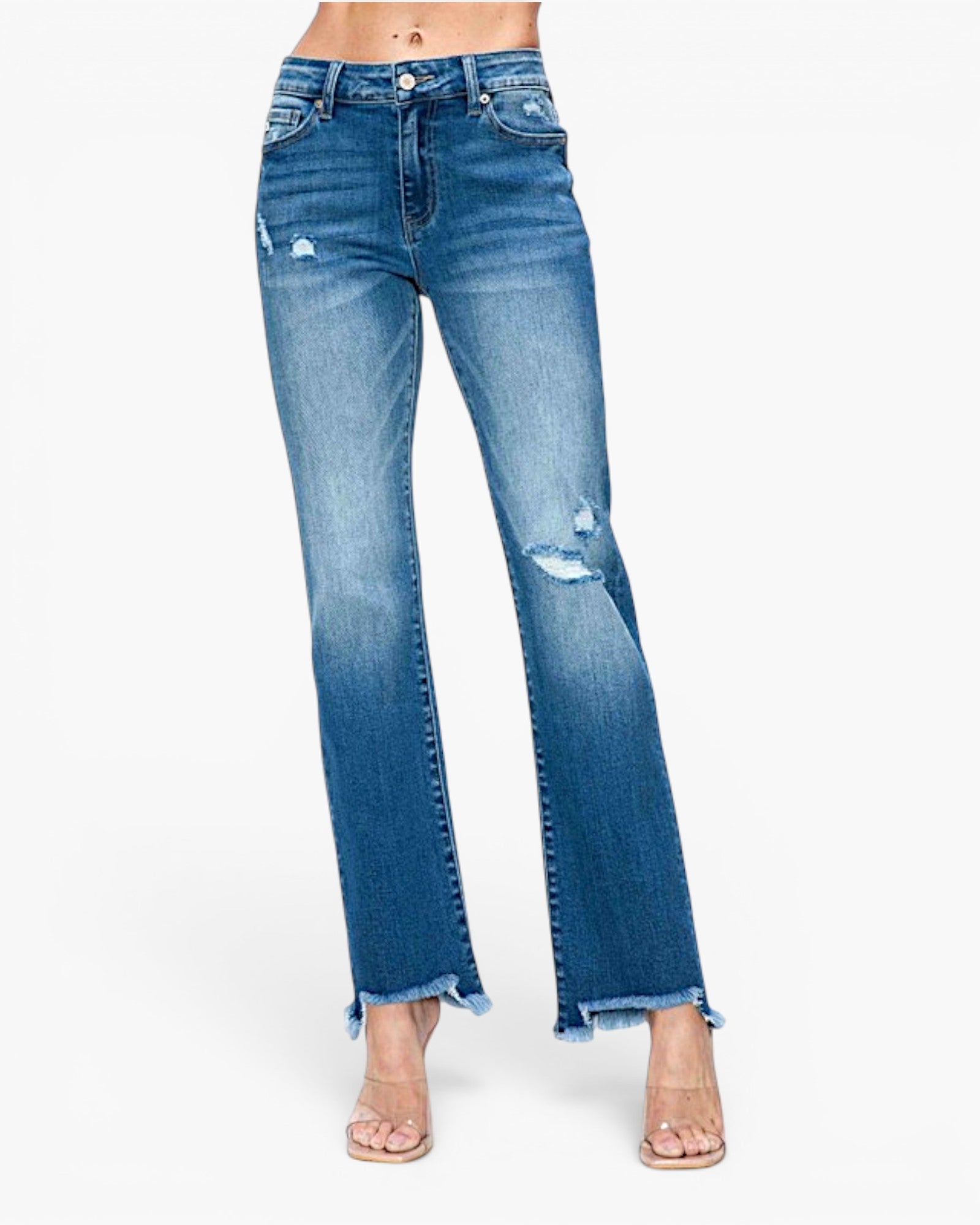 A model is wearing a pair of medium wash blue denim jeans with distressed details and frayed hems, paired with clear high-heeled sandals against a plain white background.