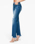 A side view of a model wearing medium wash blue denim jeans with distressed details and a raw frayed hem, paired with clear high-heeled sandals against a plain white background.