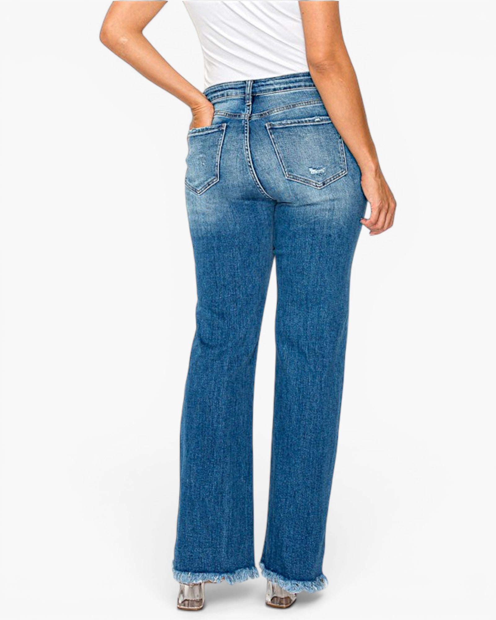 Rear view of a model wearing high-waisted, wide-leg blue denim jeans with a frayed hem, paired with a white top and metallic heels, against a plain white background.