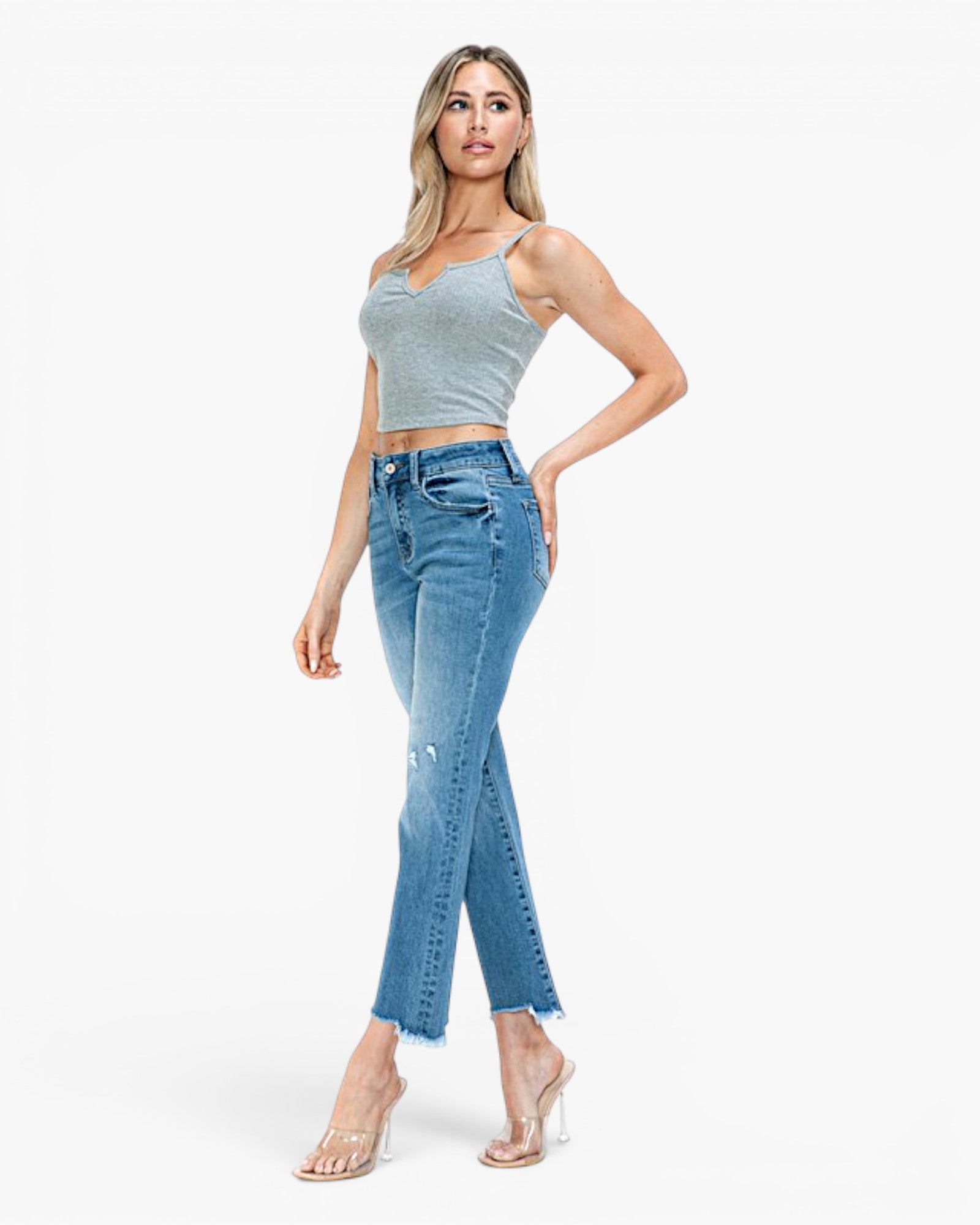 A woman modeling a pair of high-waisted, light blue cropped jeans with side cutouts, paired with a light gray crop top and clear high-heeled sandals, against a plain white studio background.