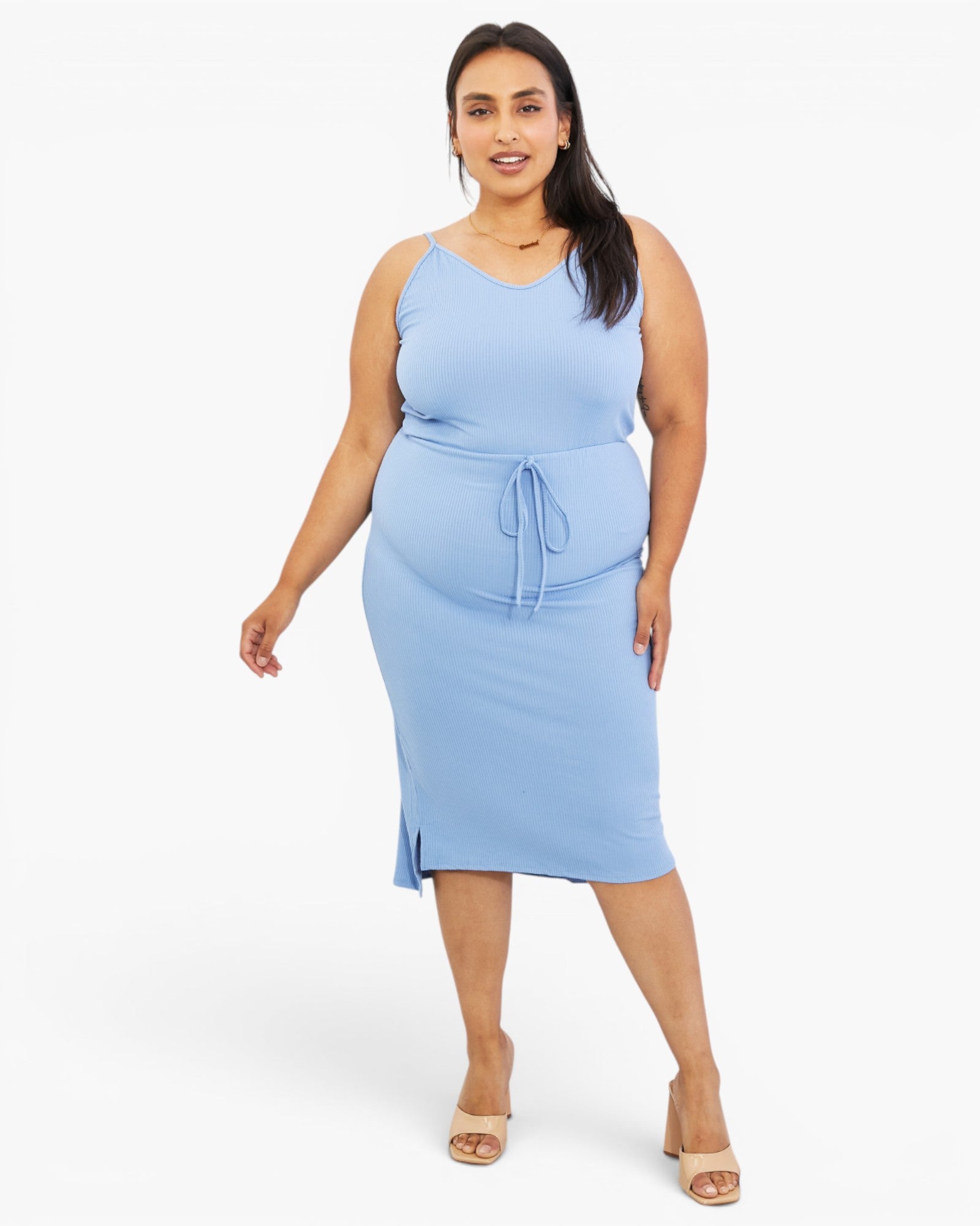 A woman modeling a light blue sleeveless midi dress with a tie waist, photographed against a plain light gray studio background.