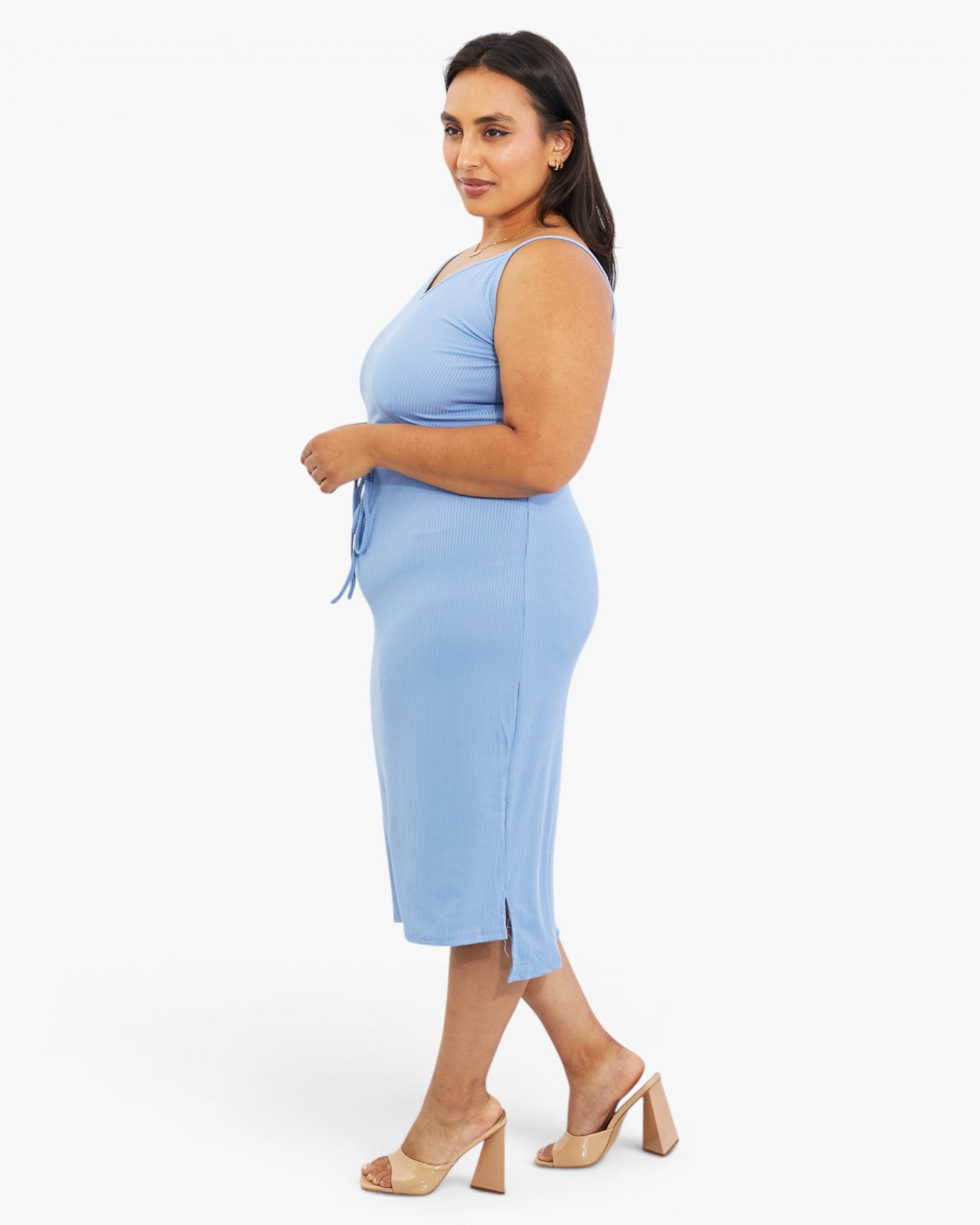 A woman modeling a light blue sleeveless midi dress with a side tie detail, paired with beige block-heel sandals, against a plain white studio background.