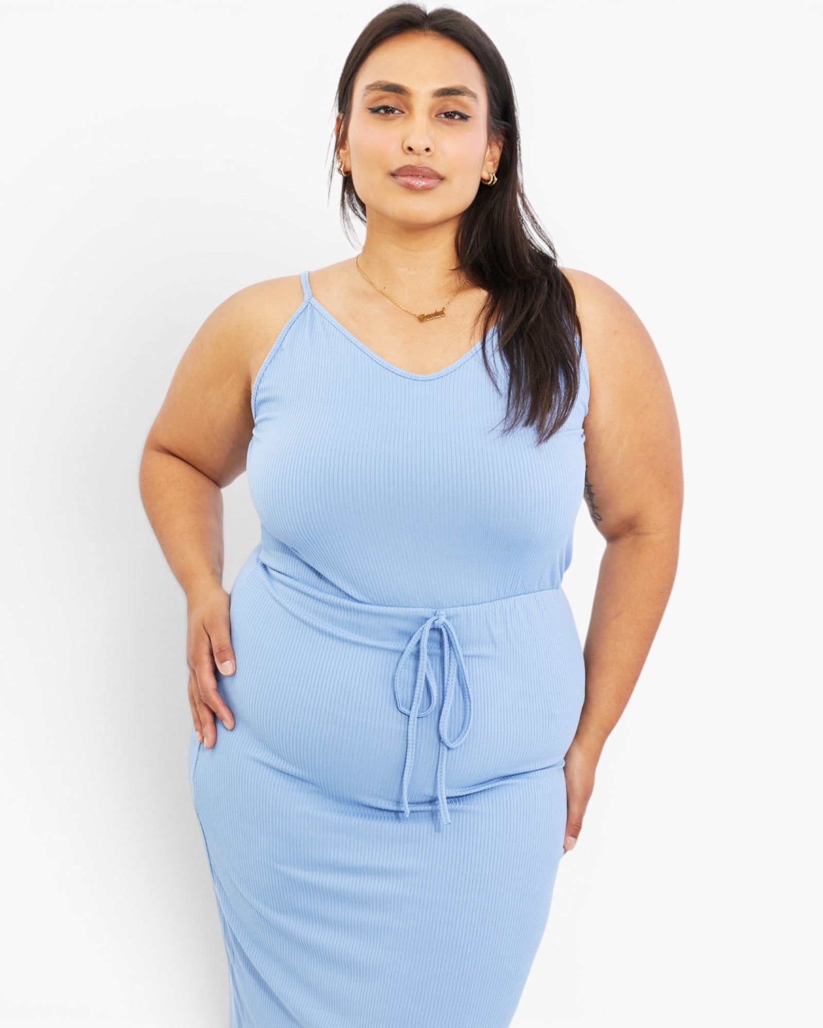 A plus-size woman modeling a light blue ribbed camisole dress with a tie-waist detail, set against a plain white studio background.