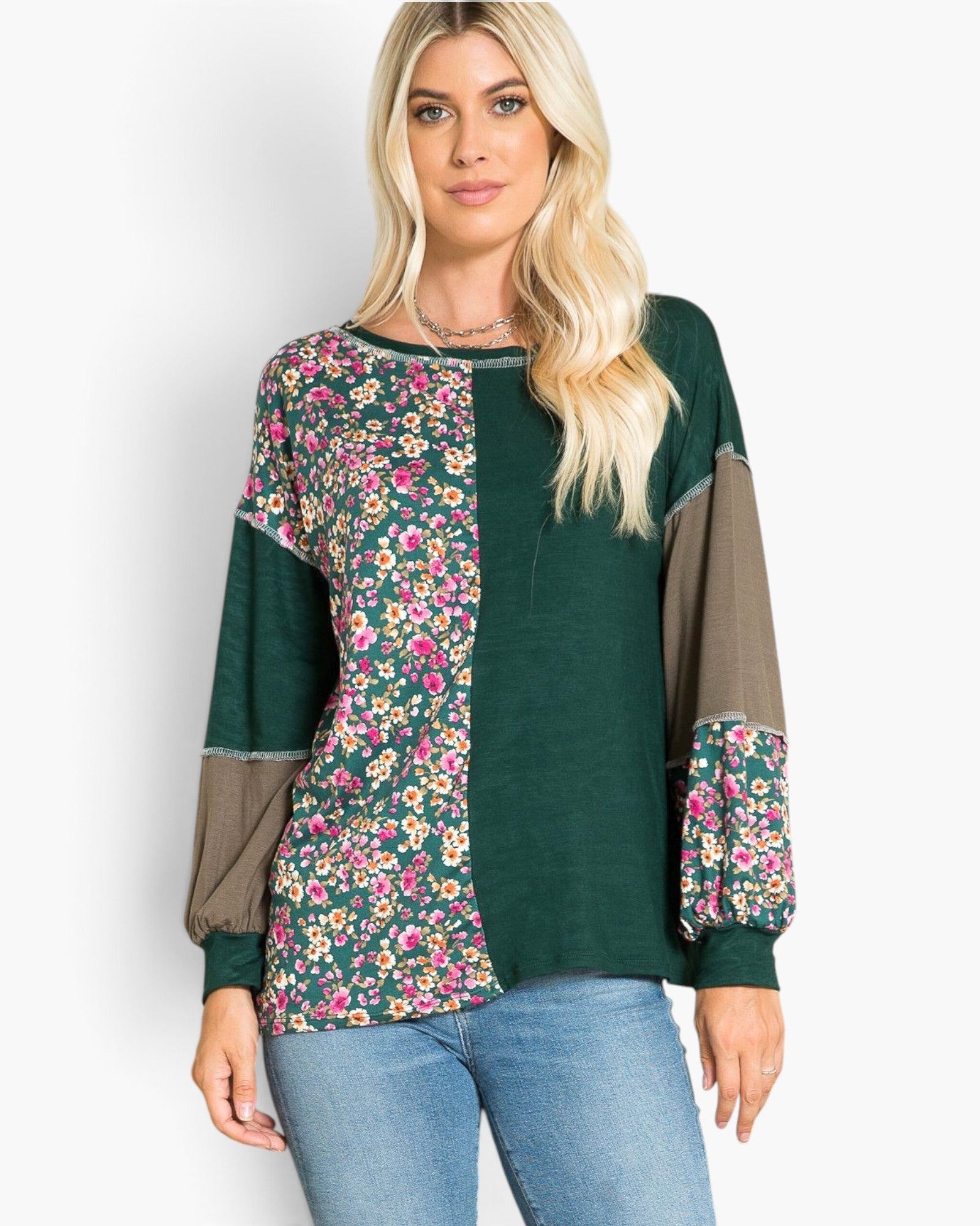 A woman modeling a long-sleeve patchwork top with a dark green base, a floral print panel on the left side, solid taupe-brown sleeves, and blue jeans against a light gray background.