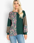 A woman modeling a long-sleeve patchwork top with a dark green base, a floral print panel on the left side, solid taupe-brown sleeves, and blue jeans against a light gray background.