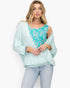 A woman modeling a light mint green draped cardigan over a turquoise floral top, paired with light-wash jeans, against a plain white background.