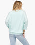 A woman shown from the back wearing a light mint green long-sleeve top and light blue jeans against a plain white background.