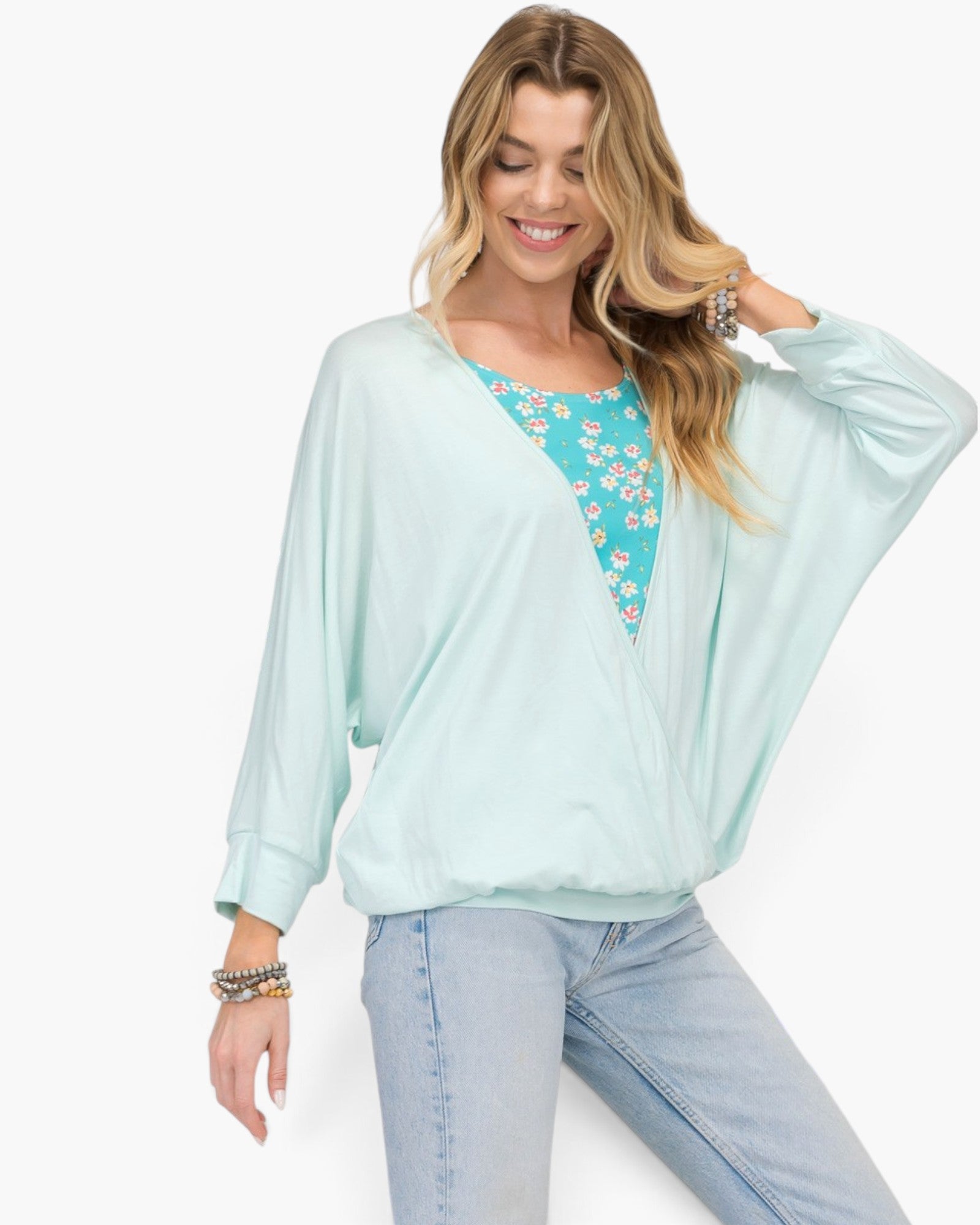 A woman wearing a light mint green long-sleeve flutter-front top over a turquoise floral tank top and light-wash jeans, posed against a clean white background.