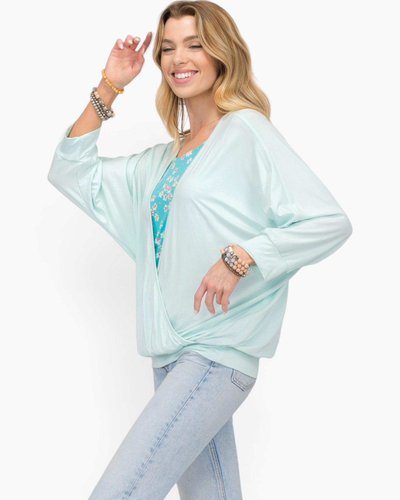 A woman modeling a light mint green kimono-style cardigan over a turquoise floral top and light blue jeans, accessorized with bracelets, against a plain white background.