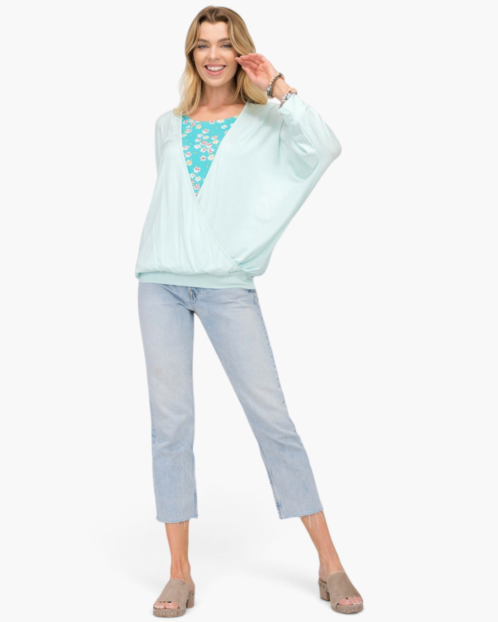 A woman modeling a light mint green wrap-style long-sleeve top over a turquoise floral top, paired with light-wash cropped jeans and beige heeled sandals, against a plain white studio background.