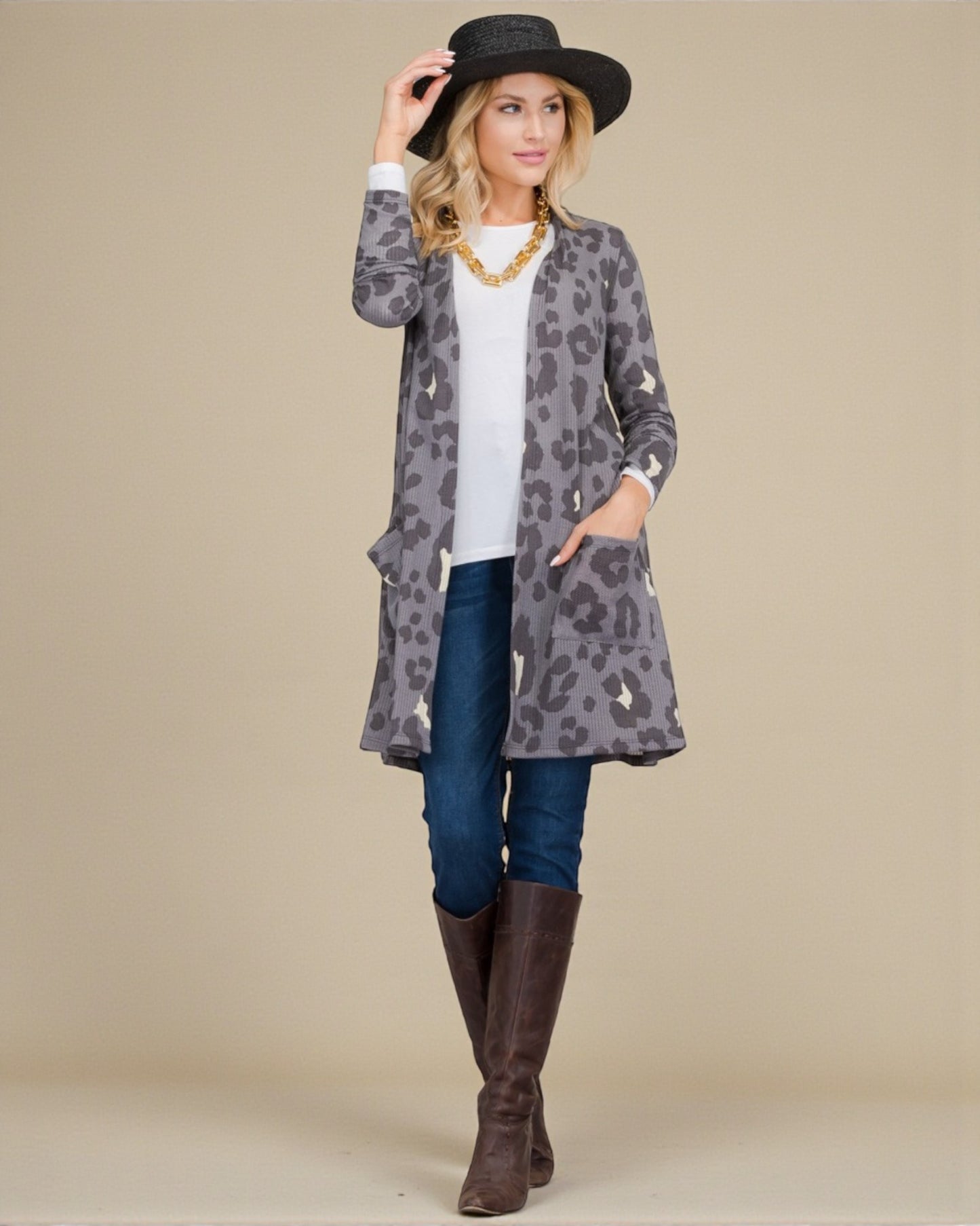 Woman wearing a gray leopard print cardigan, blue jeans, and brown boots against a beige background