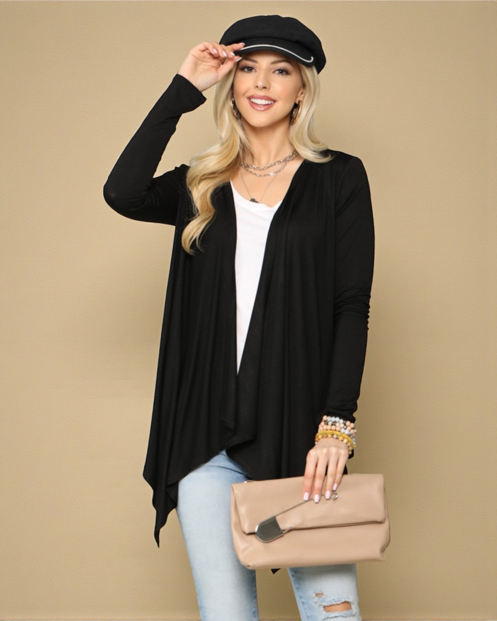 Woman wearing a black cardigan, white top, light blue jeans, and holding a beige clutch against a beige background.