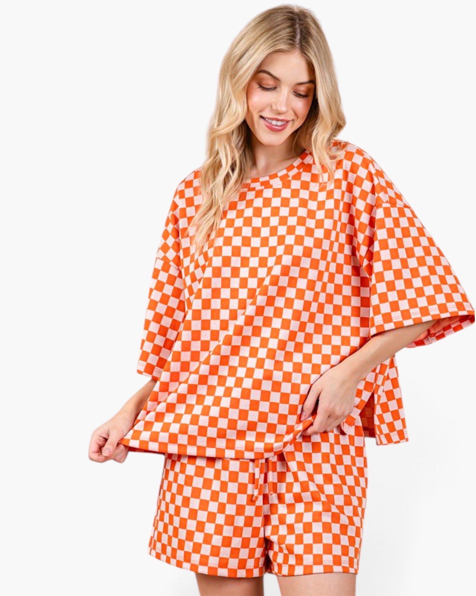 A woman modeling a matching orange and white checkerboard two-piece outfit consisting of a loose short-sleeve top and coordinating shorts, against a plain white studio background.