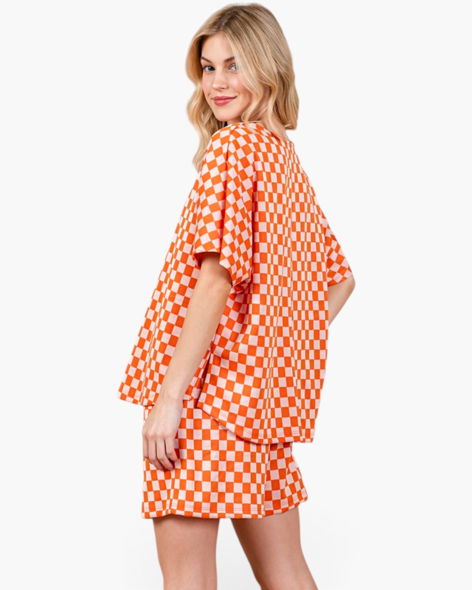 A woman modeling a bright orange and white checkerboard short-sleeve top and matching shorts against a plain light gray studio background.