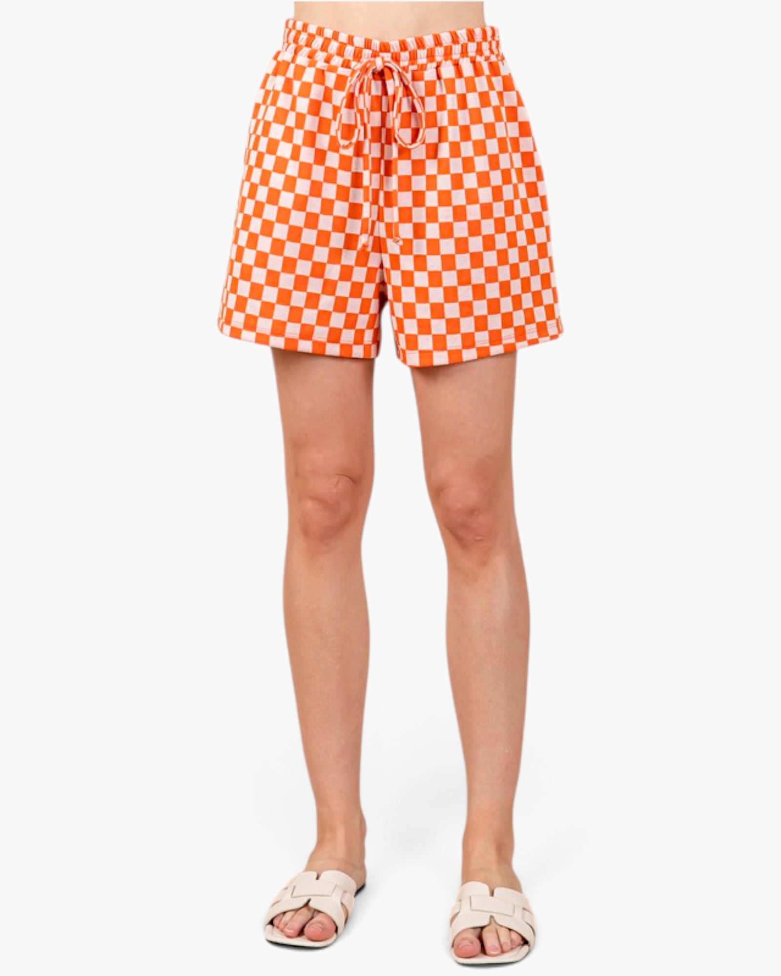 A person wearing orange and white checkered shorts with a drawstring waist, paired with light beige slide sandals, shown against a plain white studio background.