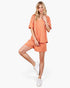 A woman modeling a two-piece lounge set featuring a short-sleeve top and matching shorts in an orange and white checkerboard pattern, paired with white sneakers, against a plain white background.