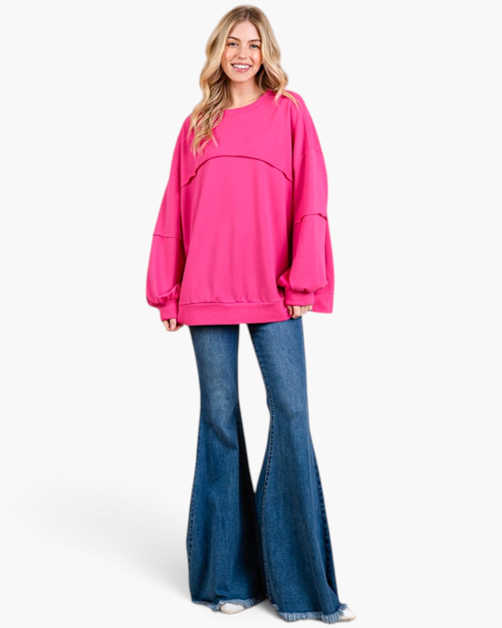 A woman modeling an oversized bright pink crewneck sweatshirt with layered long sleeves and blue flared jeans against a plain white studio background.