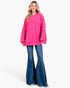 A woman modeling an oversized bright pink crewneck sweatshirt with layered long sleeves and blue flared jeans against a plain white studio background.
