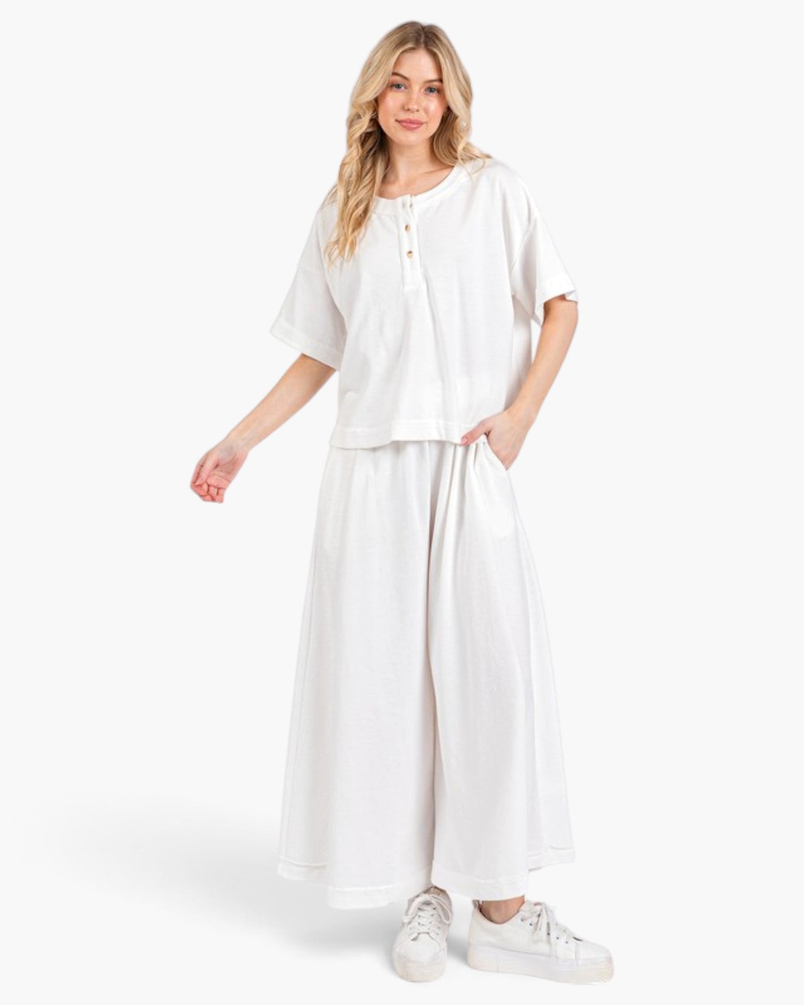 A woman modeling a white two-piece linen set consisting of a short-sleeve henley top and matching wide-leg pants, paired with white sneakers, against a light gray studio background.