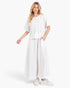 A woman modeling a white two-piece linen set consisting of a short-sleeve henley top and matching wide-leg pants, paired with white sneakers, against a light gray studio background.