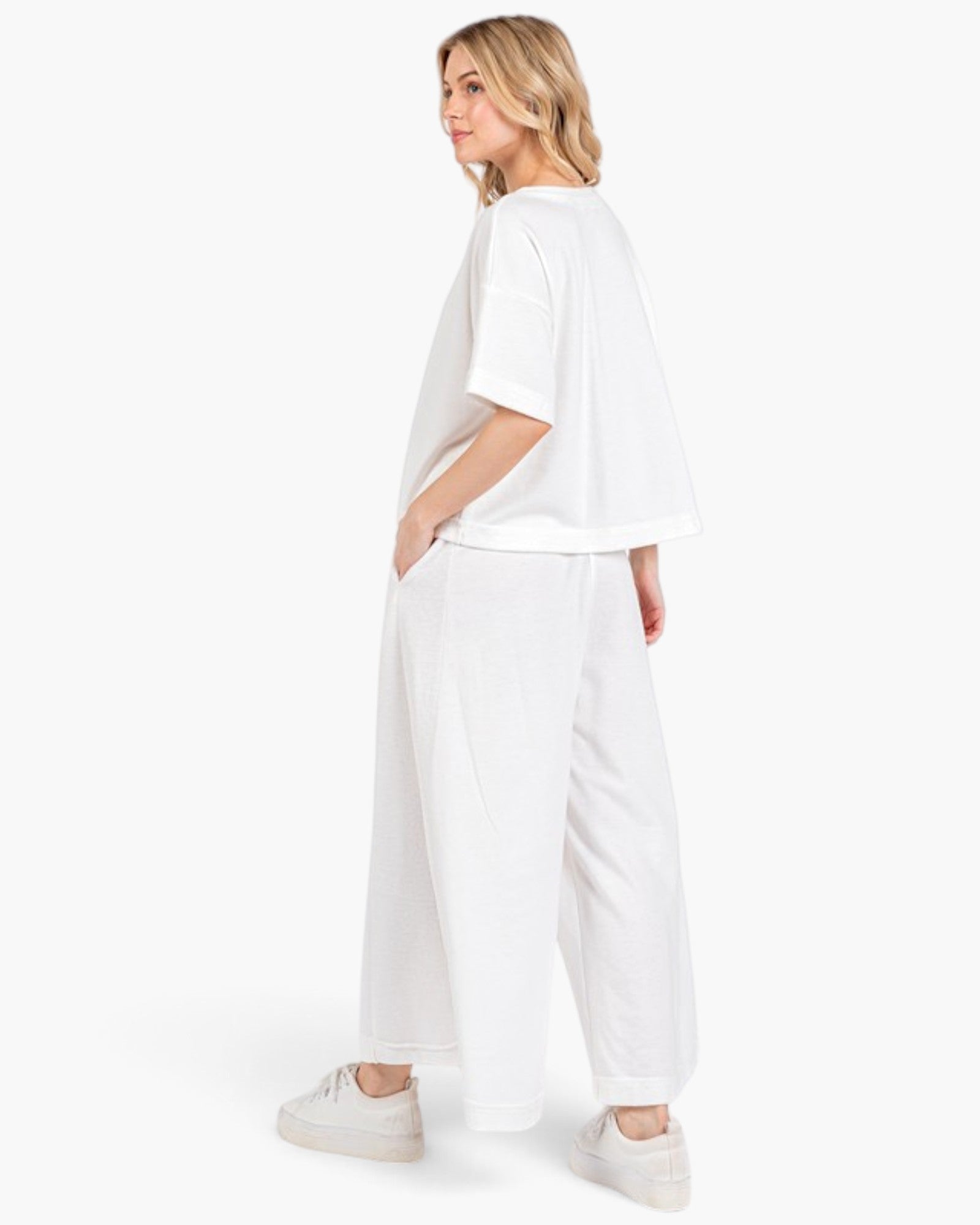 A woman modeling a white oversized short-sleeve t-shirt and matching white wide-leg pants, paired with white sneakers, shown from a side-back angle against a plain white studio background.