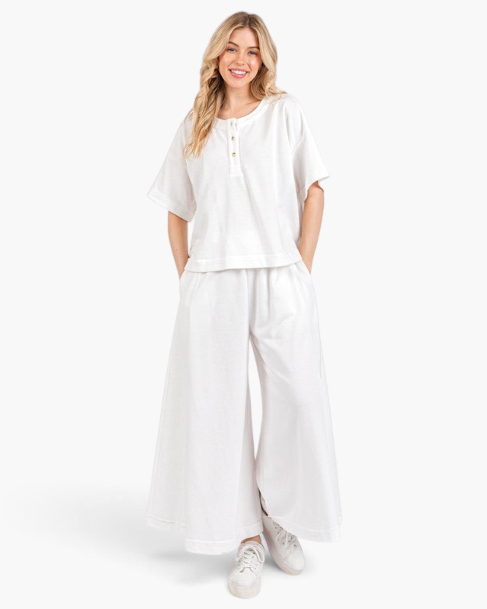 A woman modeling a white two-piece lounge set consisting of a short-sleeve henley top and matching wide-leg pants, paired with white sneakers against a light gray studio background.