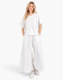 A woman modeling a white two-piece lounge set consisting of a short-sleeve henley top and matching wide-leg pants, paired with white sneakers against a light gray studio background.