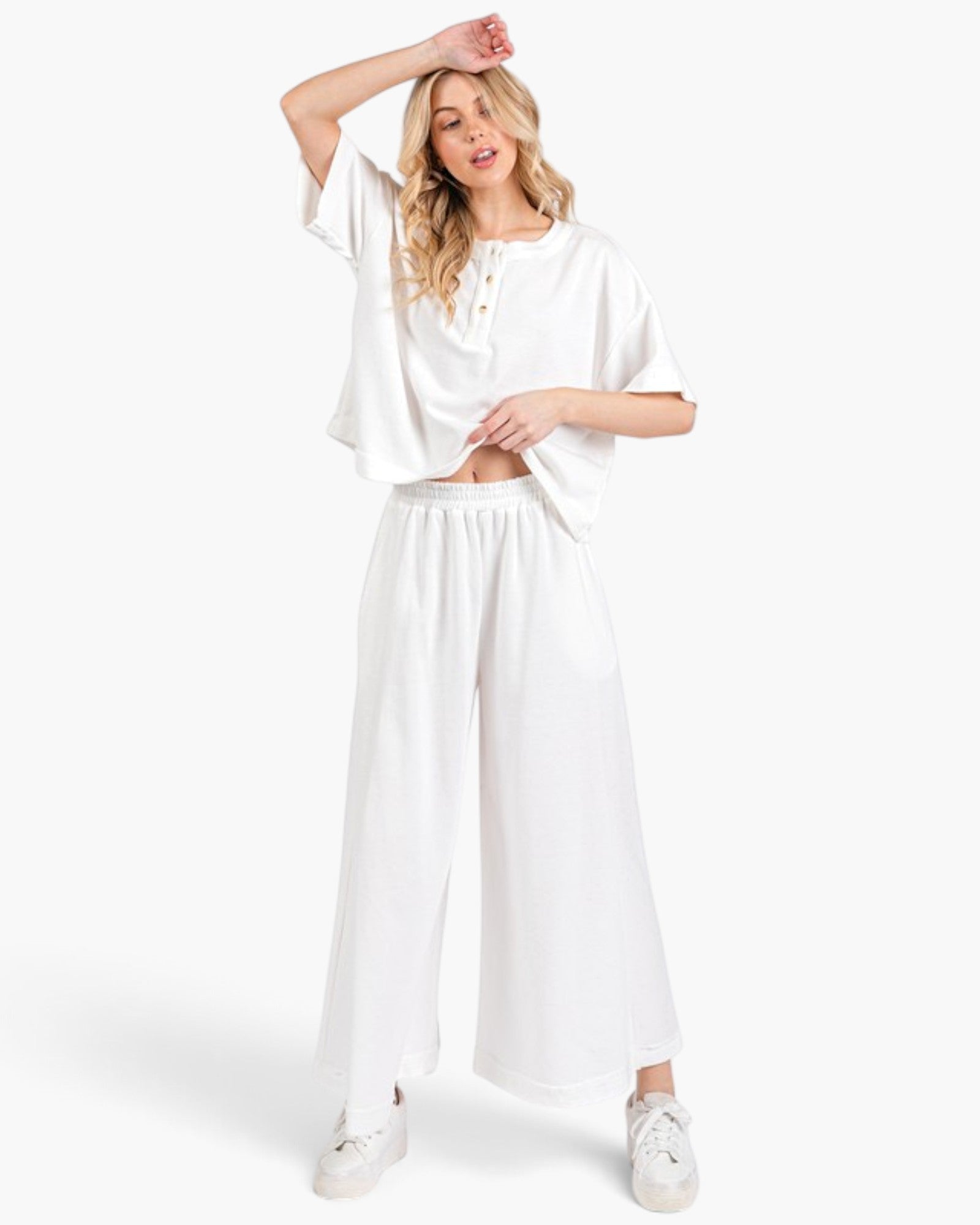 A woman modeling a white two-piece lounge set consisting of a short-sleeve cropped henley top and matching wide-leg pants, paired with white sneakers against a light gray studio background.