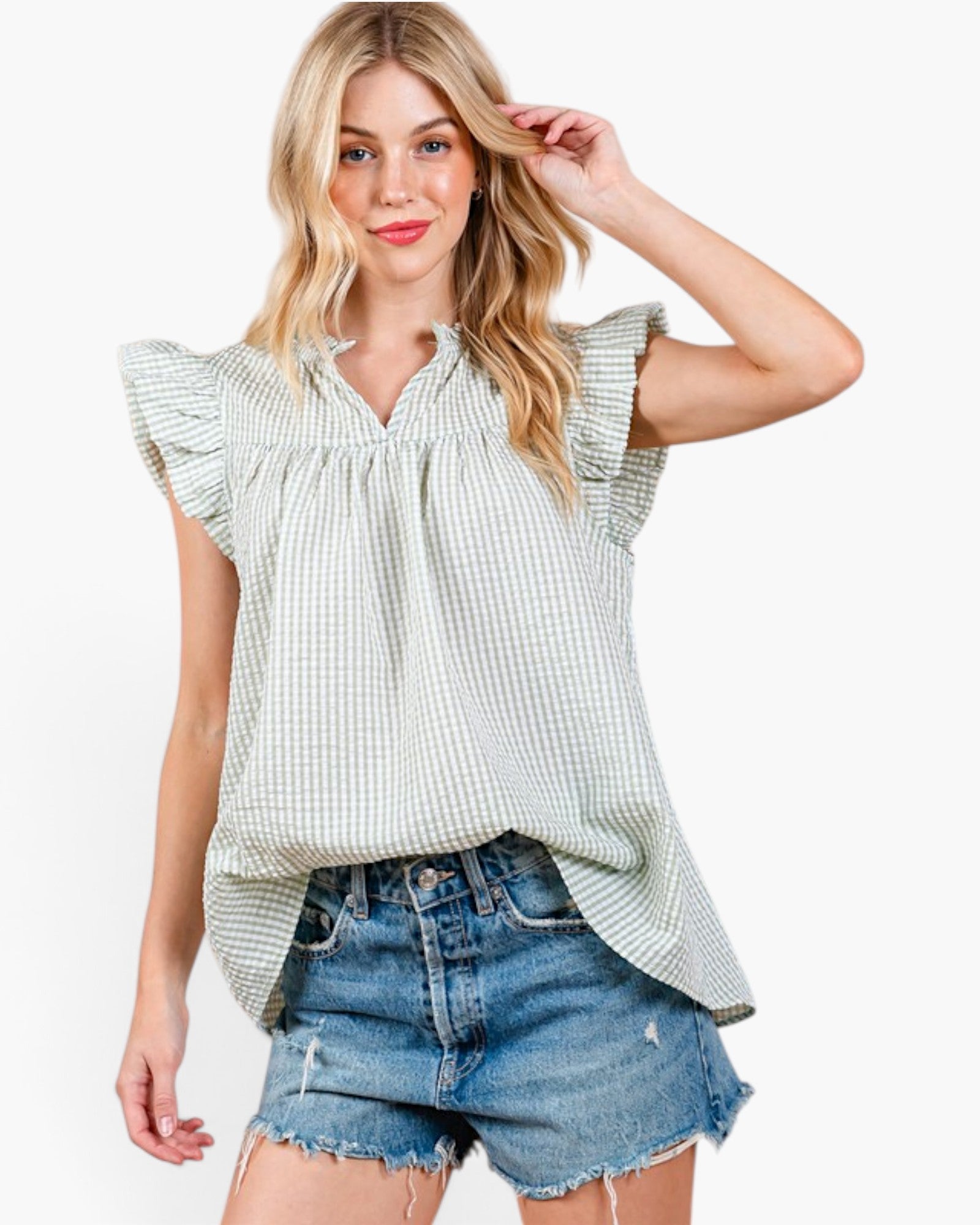 A woman modeling a light green and white gingham sleeveless blouse with ruffled cap sleeves and a smocked neckline, paired with distressed denim shorts against a plain white background.