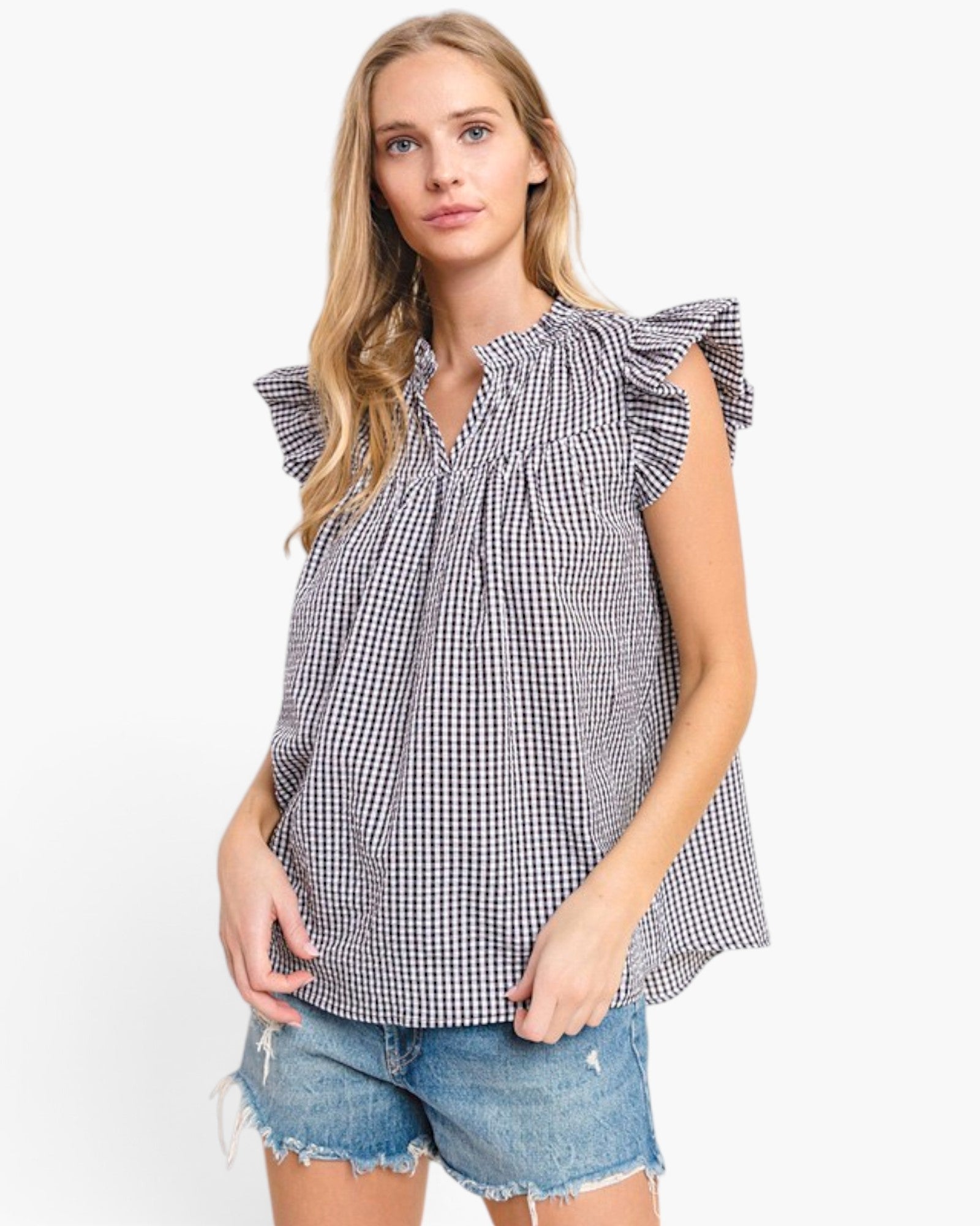 A woman modeling a sleeveless black and white gingham top with ruffled shoulder details and a smocked neckline, paired with denim shorts, against a plain white background.