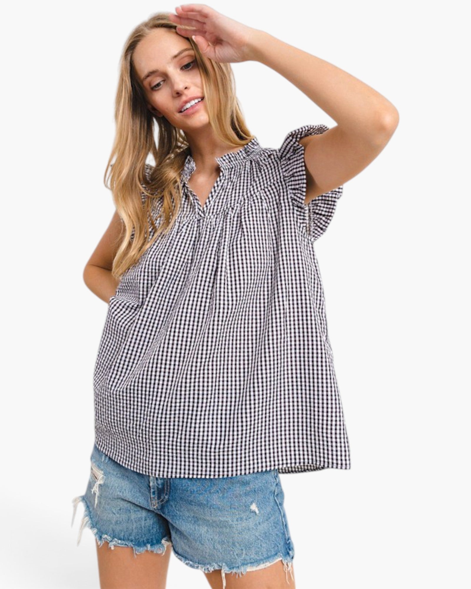 A woman modeling a black and white gingham check short-sleeve blouse with ruffled shoulders and a smocked neckline, paired with distressed denim shorts, against a plain white background.