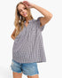 A woman modeling a black and white gingham check short-sleeve blouse with ruffled shoulders and a smocked neckline, paired with distressed denim shorts, against a plain white background.
