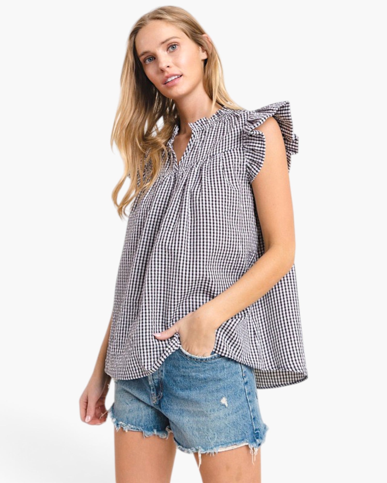A woman modeling a sleeveless black and white gingham blouse with ruffled shoulders and a smocked neckline, paired with denim shorts, against a plain white studio background.