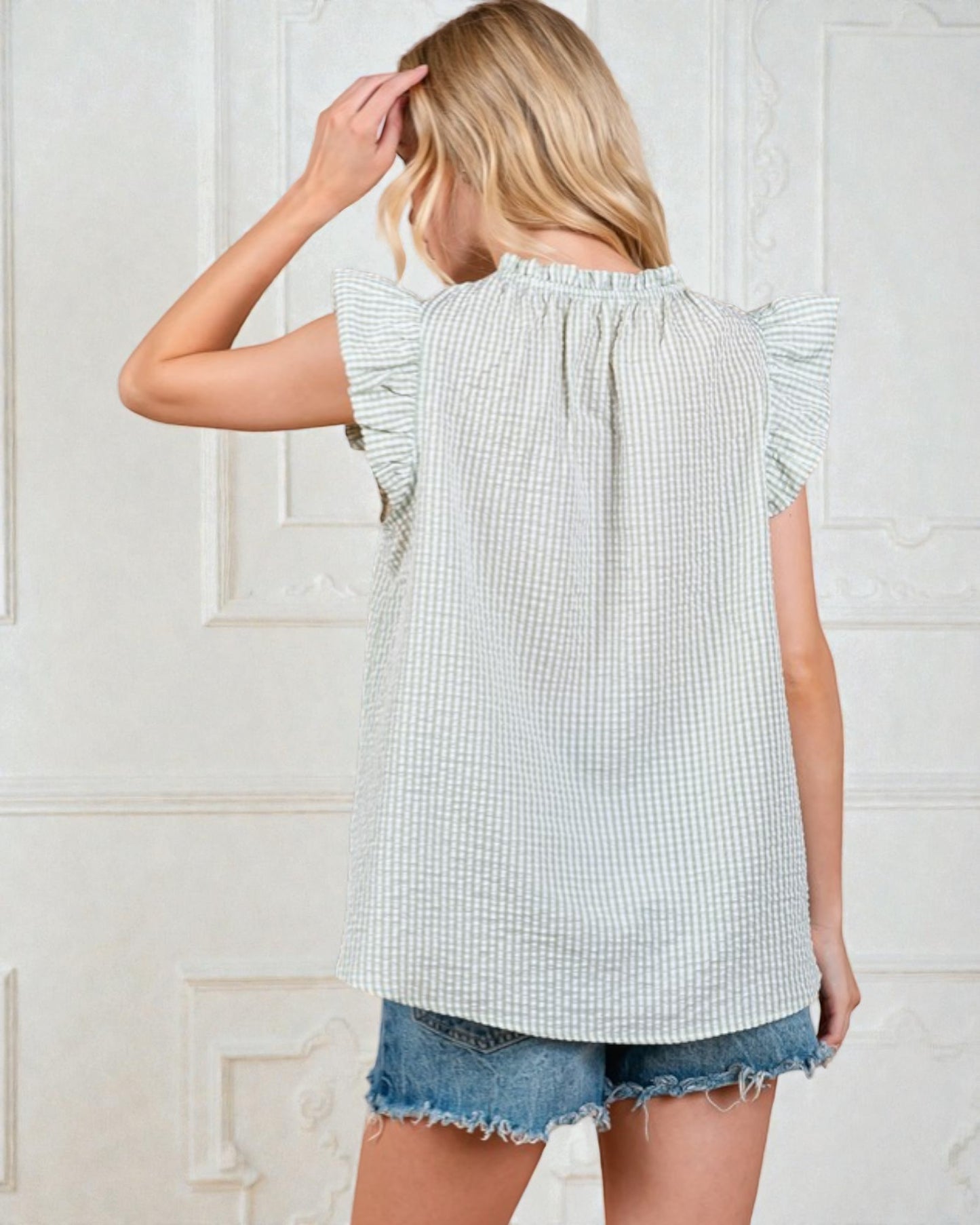 Woman wearing a light blue checkered top and denim shorts against a white wall.