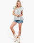 A woman modeling a light sage green and white vertically striped ruffled sleeveless top, paired with denim shorts and cream ankle boots, against a plain white background.