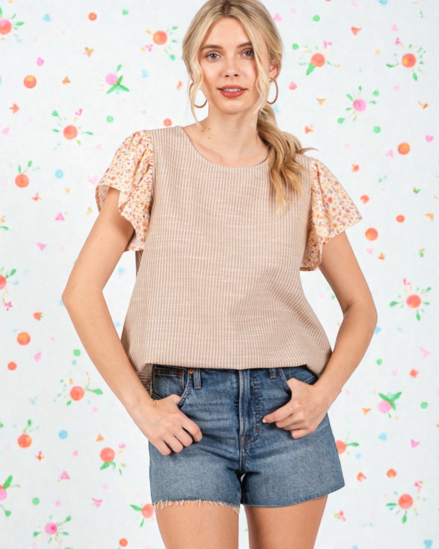 Woman wearing a beige ribbed top with floral sleeves and denim shorts against a colorful abstract background