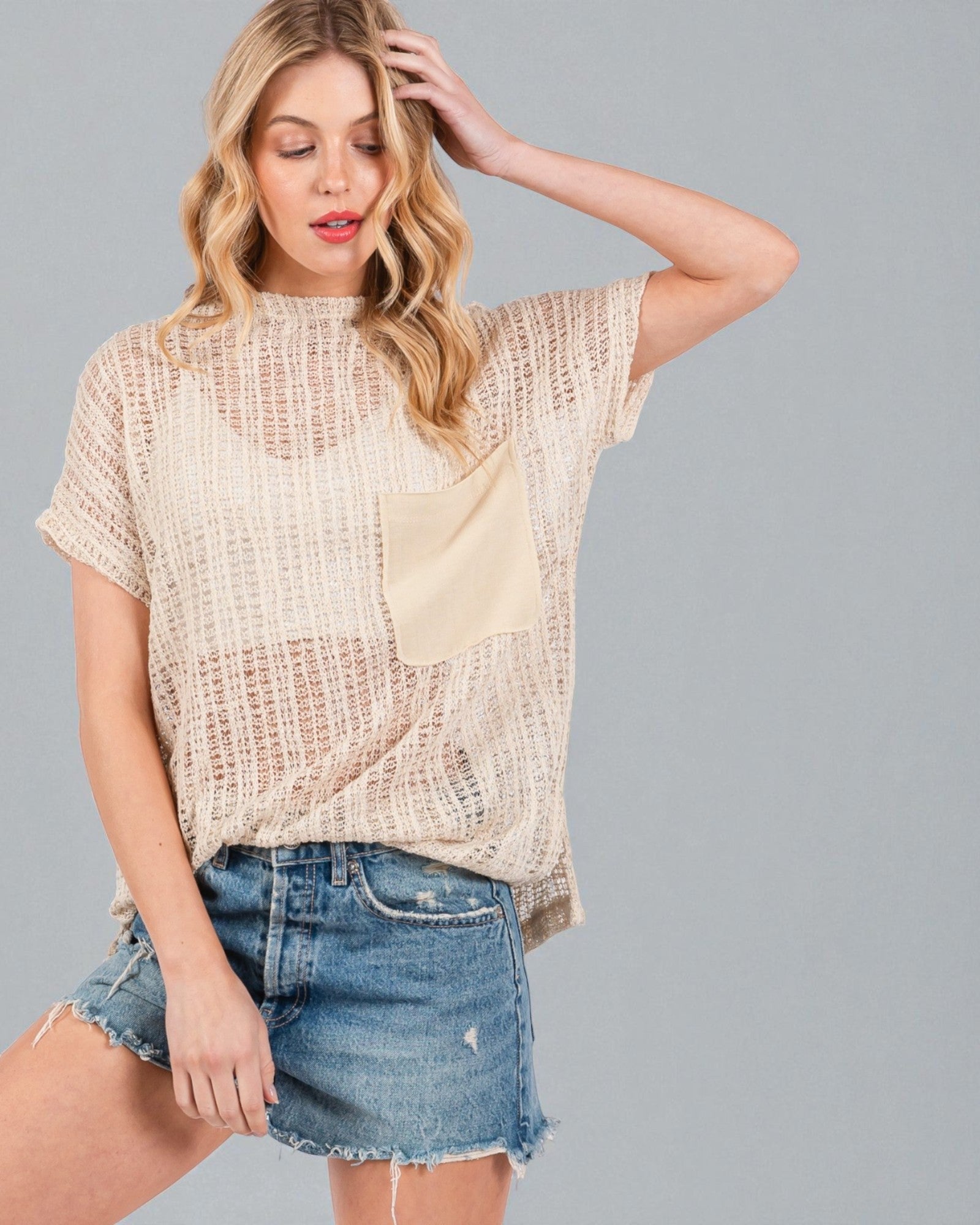 Woman wearing a beige mesh top and denim shorts on a gray background