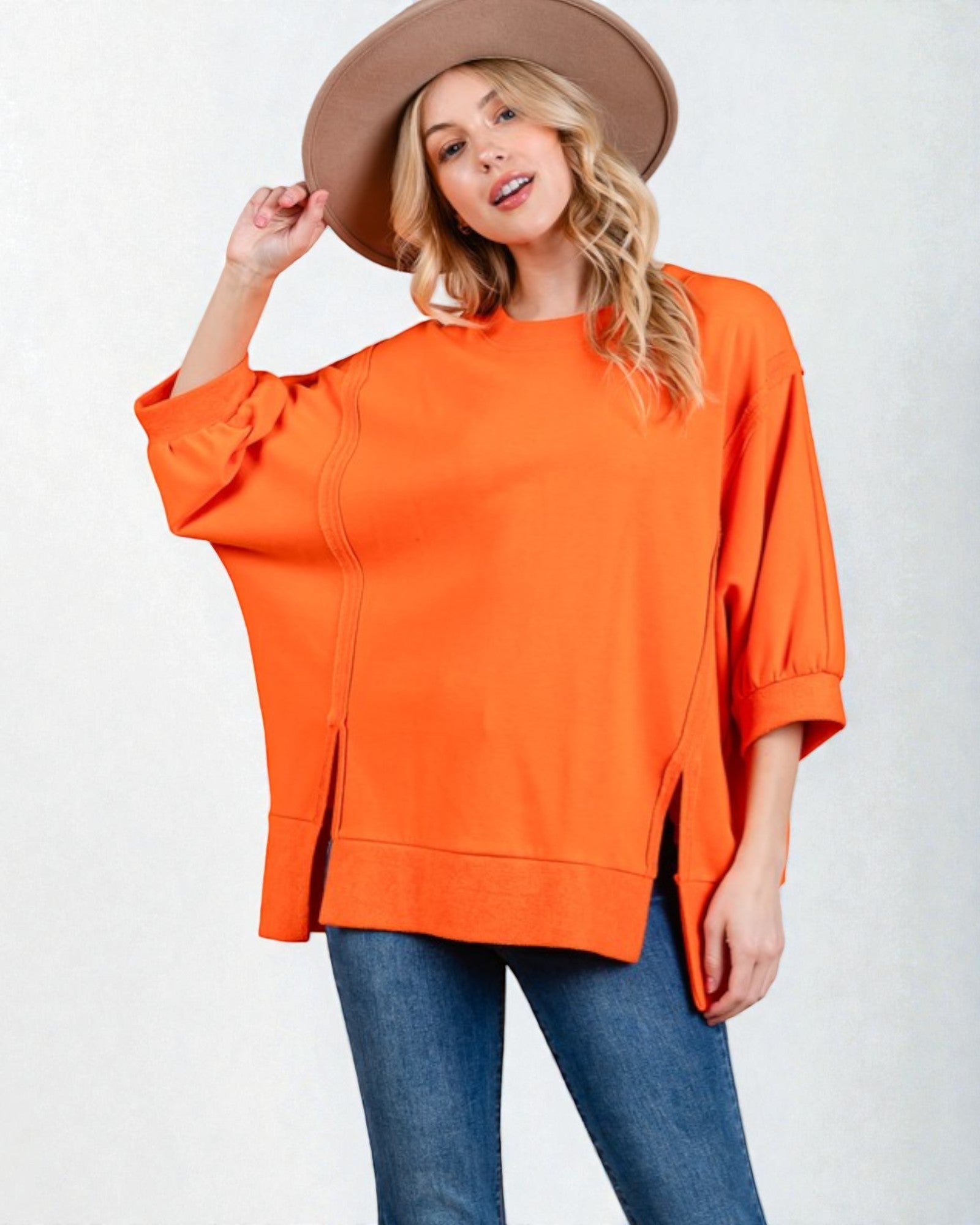 Woman wearing an orange oversized top and blue jeans with a beige hat on a white background