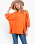 A woman wearing a bright orange oversized long-sleeve top with side slits, blue jeans, and a brown wide-brim hat, photographed against a clean white studio background.