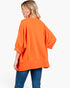 A woman shown from the back wearing a bright orange oversized top with three-quarter sleeves and blue jeans, photographed against a plain light gray studio background.