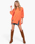 A woman modeling a loose-fitting orange long-sleeve top with side slits, paired with denim shorts and brown knee-high boots, against a plain light gray studio background.