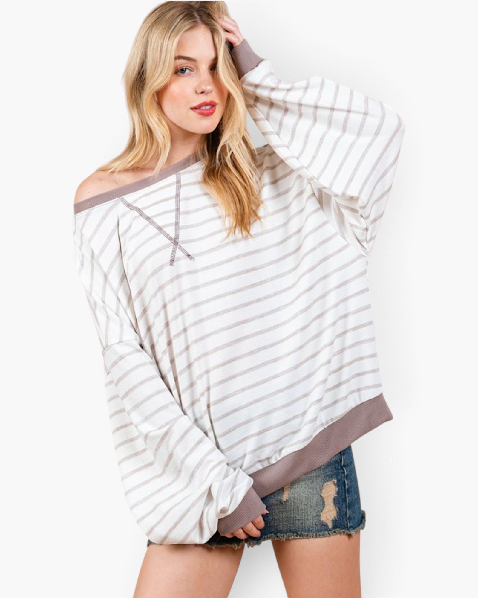 A woman modeling a loose off-the-shoulder long-sleeve top with thin gray horizontal stripes on an off-white base, paired with distressed denim shorts against a plain white background.