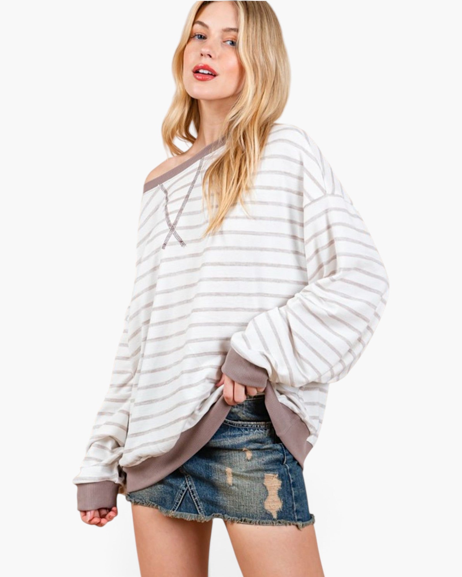 A woman modeling a long-sleeve, off-the-shoulder striped top with a taupe trim, paired with distressed denim shorts, against a plain white studio background.