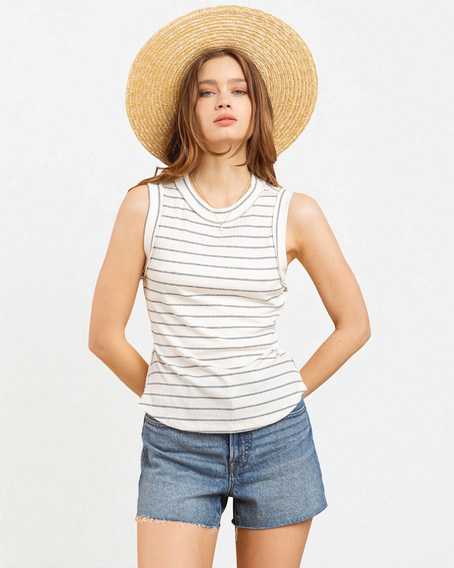 Woman wearing a striped tank top, denim shorts, and a straw hat on a light gray background